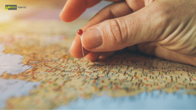 A close-up of a hand placing a red pin on a map, highlighting a specific location. The map is filled with detailed roads and city names, suggesting travel planning