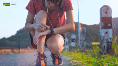 A person in athletic wear, crouching on a road while checking the time on a smartwatch. The individual wears earphones and is surrounded by green grass and a distant mountain. The focus is on the smartwatch, likely tracking fitness data.
